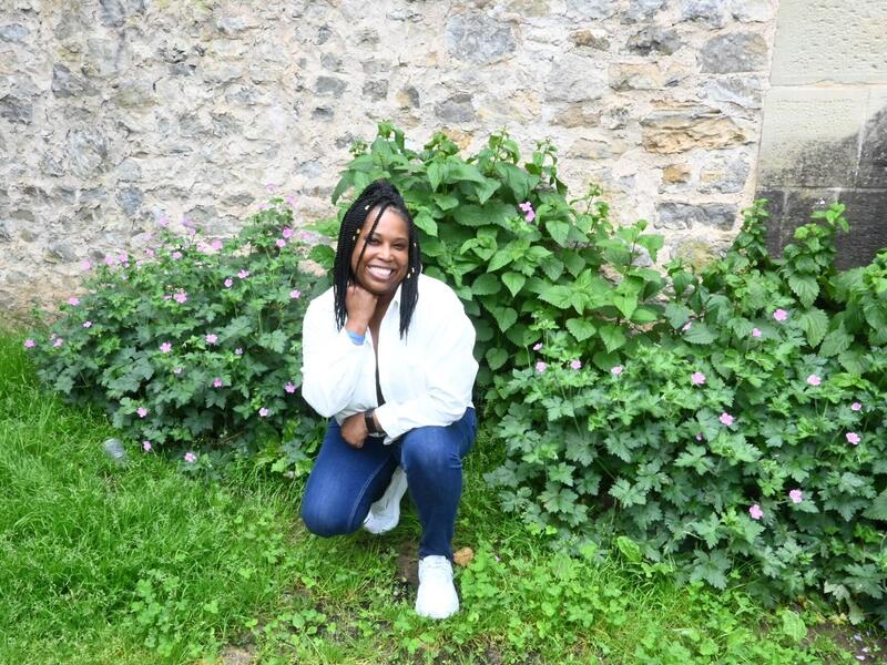 A photo of a woman kneeling in a patch of grass in front of an old gray stone wall. 