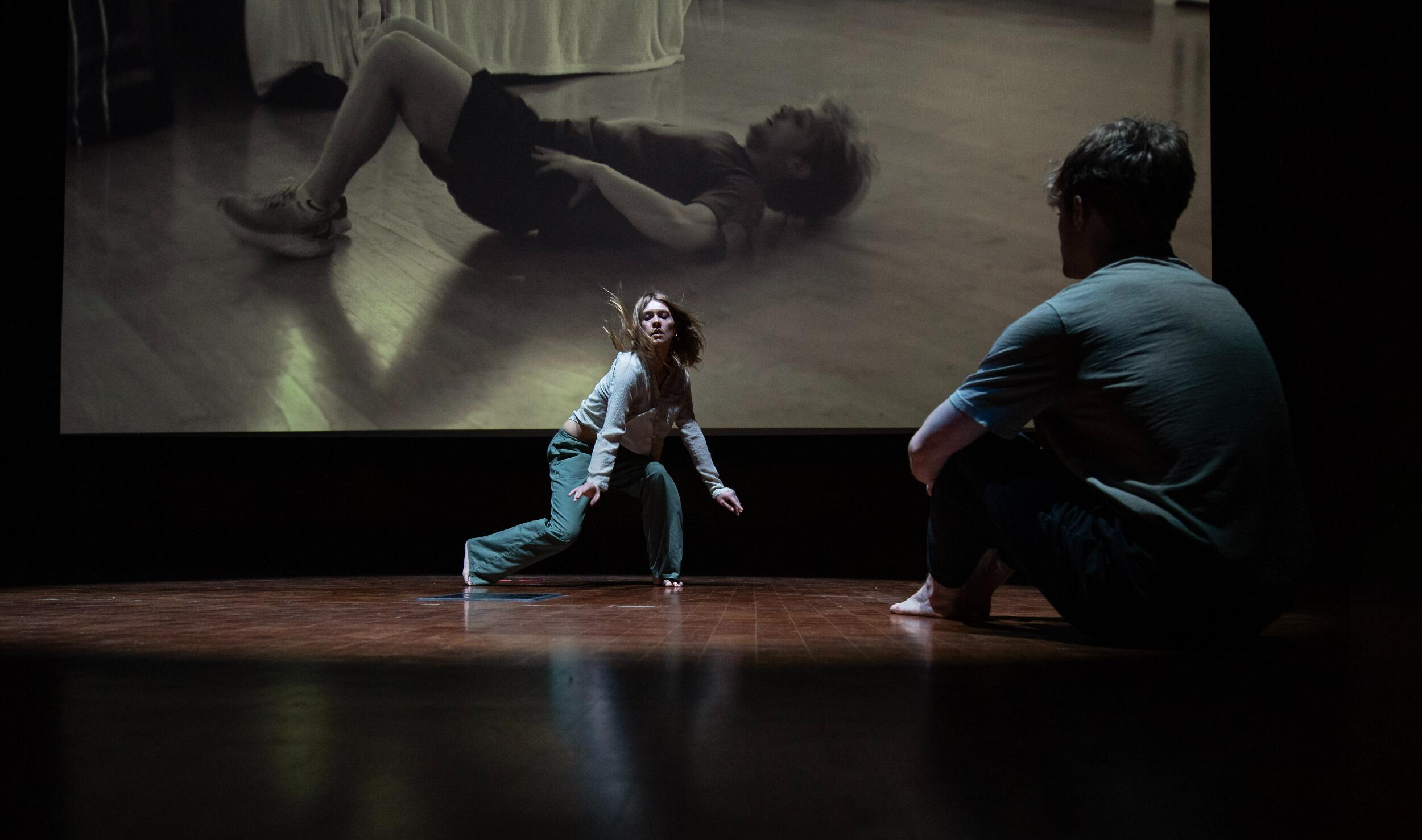 A photo of a person sitting on the floor watching another woman dance. 