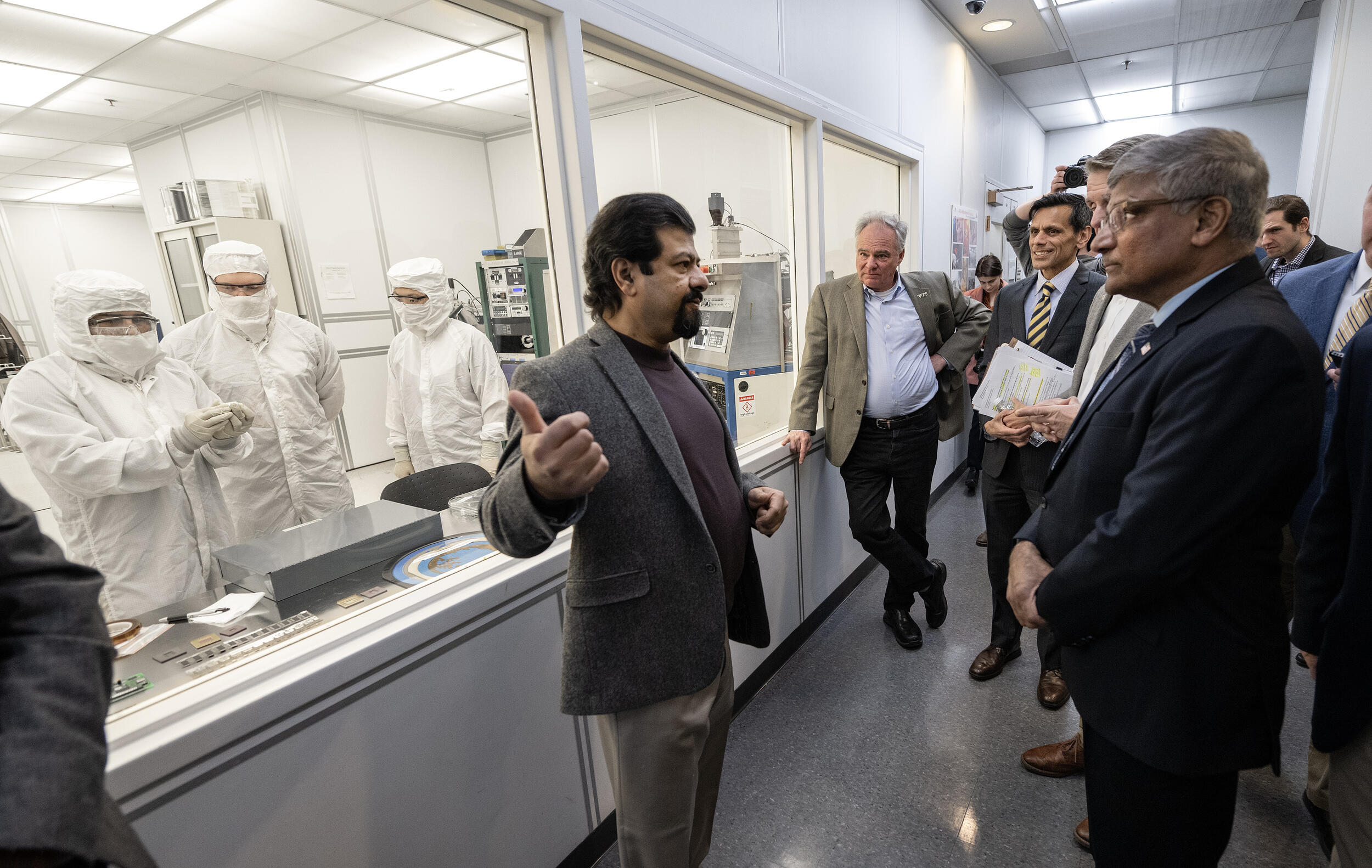 A photo of a man speaking to a group of people in front of a window showing three men in lab coats working
