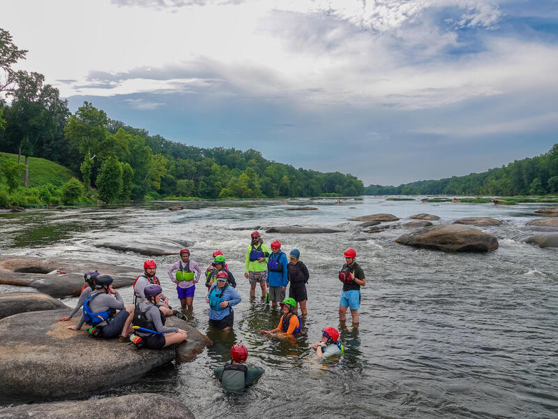 A photo of a group of people in the middle of a river. One man is standing in the middle of the group speaking. There is a circle of people around him listening. Some people are standing, and others are sitting on rocks or in the water. 