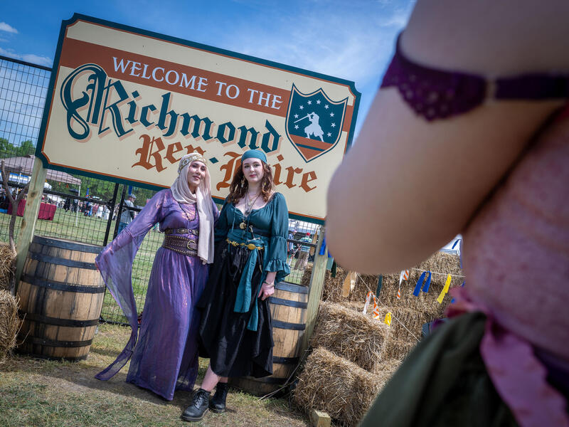 A photo of two women standing in front of a sign that says \"WELCOME TO THE Richmond Ren-Faire.\" There is a person in front of them who is taking a photo of the two. 