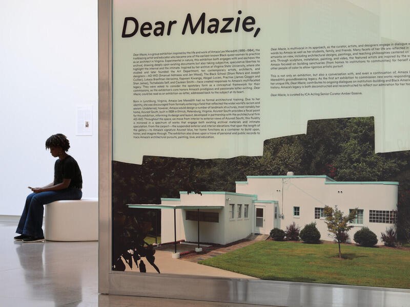 A photo of a sign that says \"Dear Mazie,\" with smaller text underneath that is illegible. There is a photo of a house underneath the text. To the left of the sign is a person sitting on a white bench.