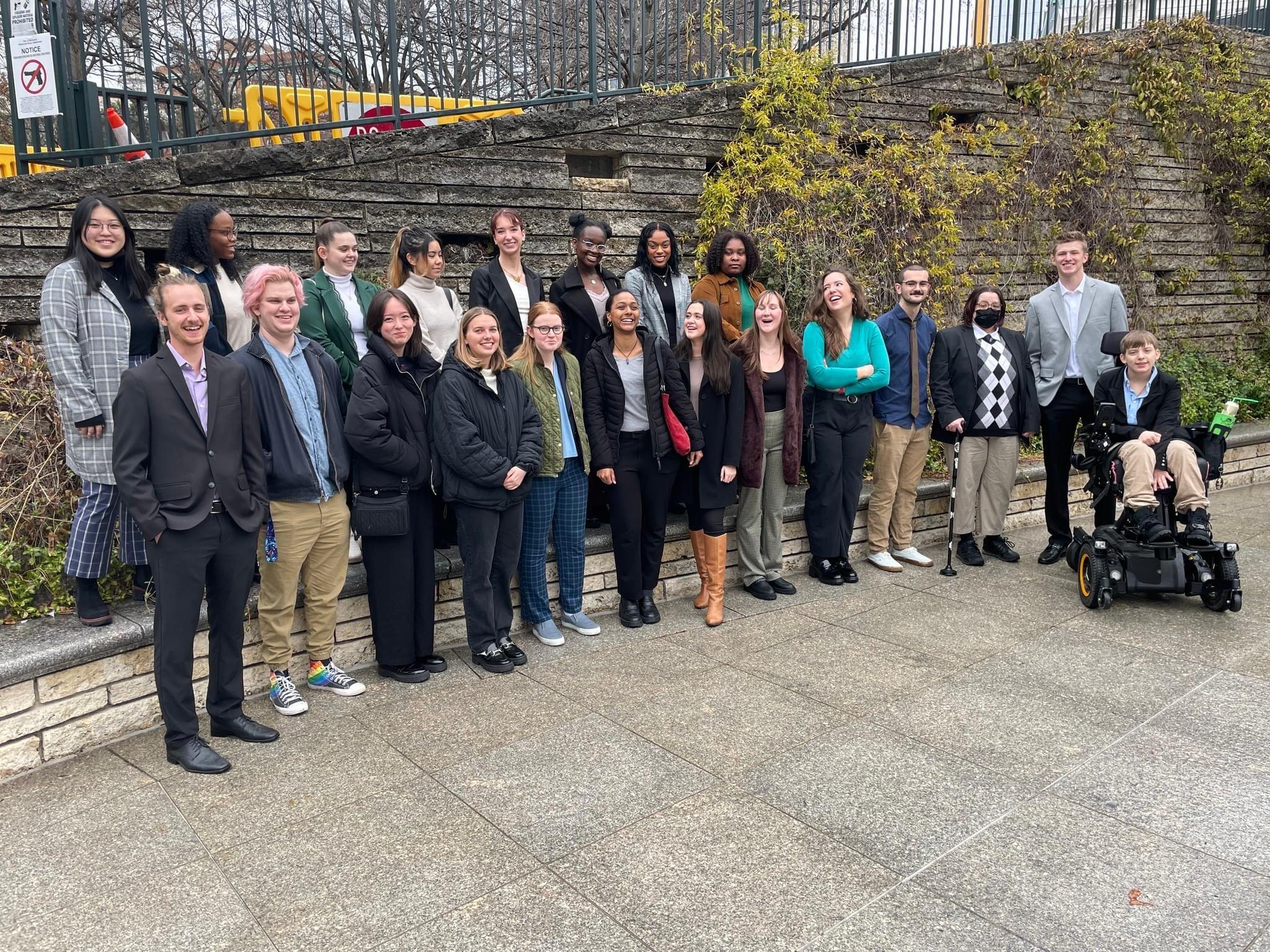 A group of 20 college students standing in front of a gray stone wall with plant vines growing up the sides of it. One student is sitting in an electric wheelchair. 