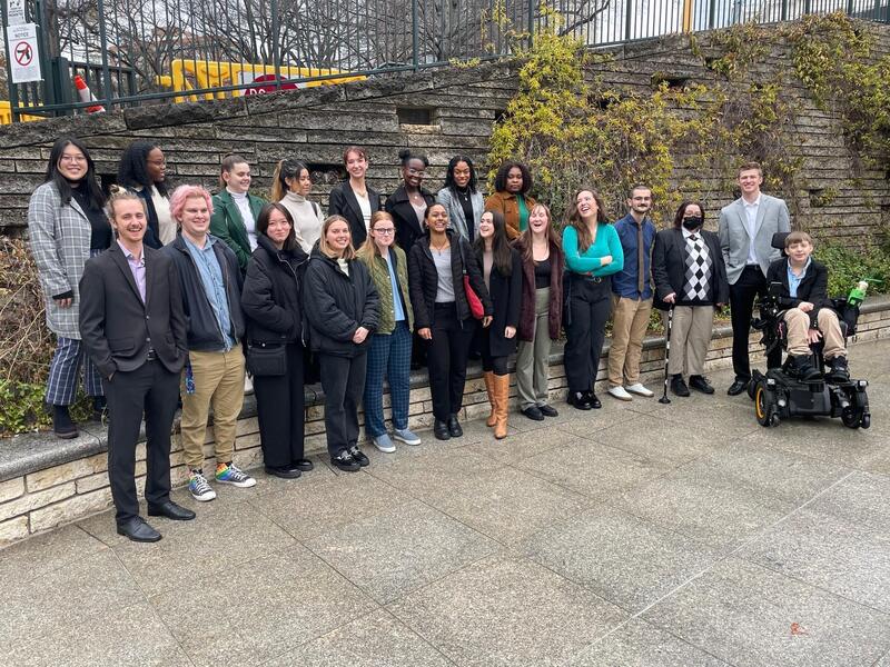 A group of 20 college students standing in front of a gray stone wall with plant vines growing up the sides of it. One student is sitting in an electric wheelchair. 