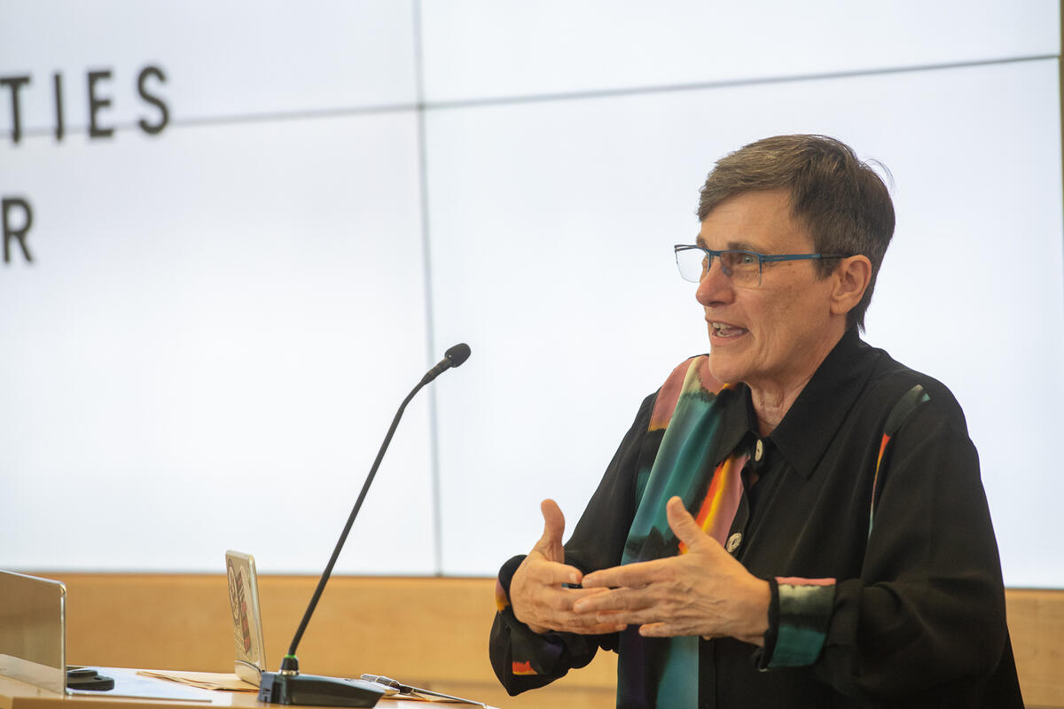 A photo of a person speaking at a podium with a microphone. The person is gesturing their hands in front of them. 