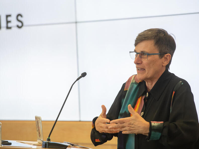 A photo of a person speaking at a podium with a microphone. The person is gesturing their hands in front of them. 