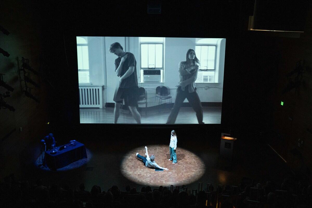A photo of two people dancing on a stage while a projection of two people dancing plays behind them. 