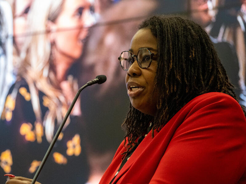 A photo of a woman from the chest up. She is facing the left and speaking into a microphone. 