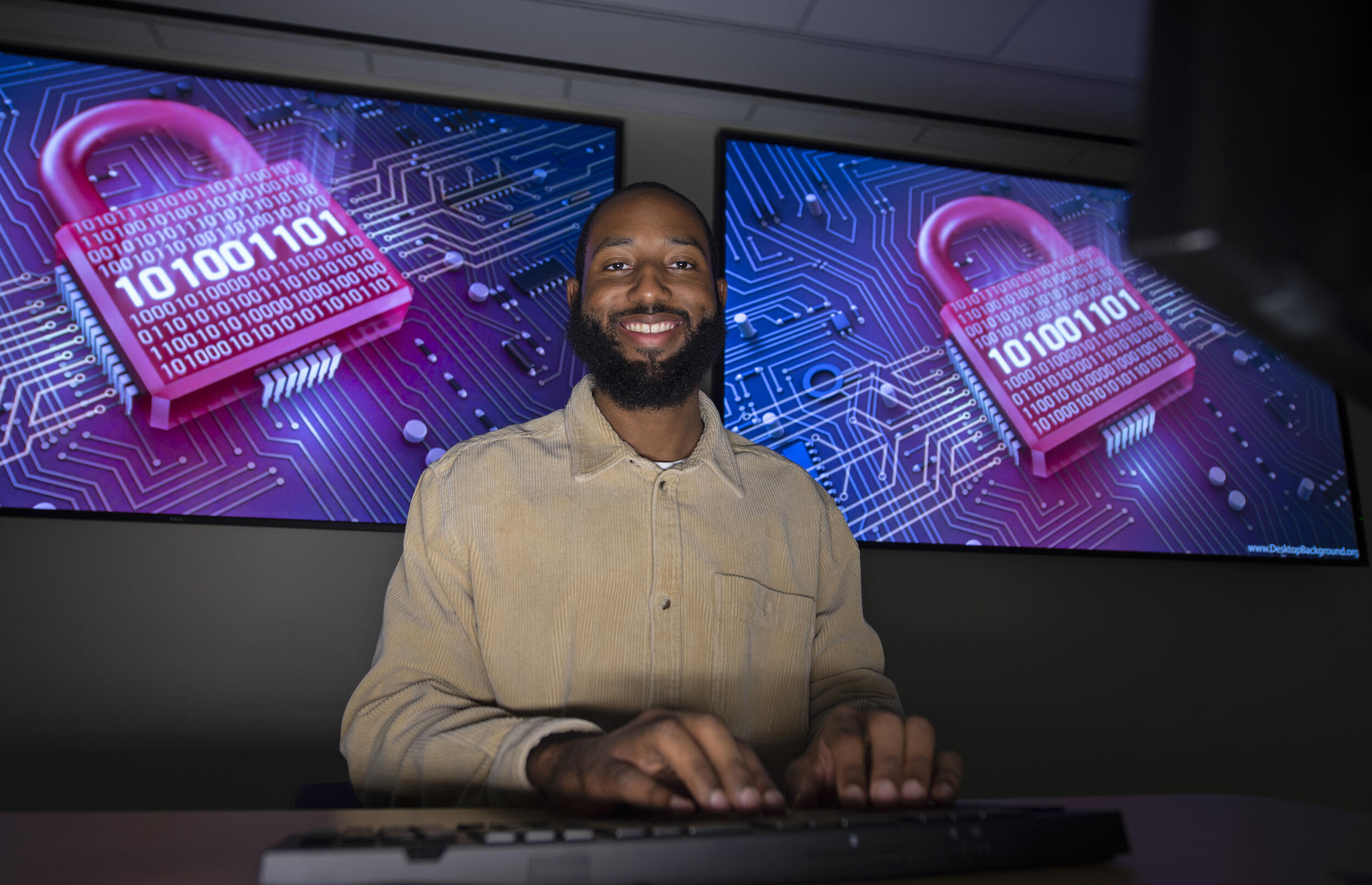 A photo of a man sitting at a desk with his hands on a keyboard. Behind him are two screens with images of locks on them. 