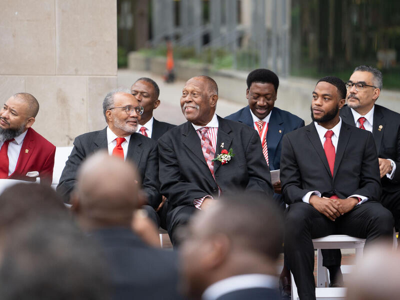 A photo of seven men sitting in front of a crowd of people in the foreground. 
