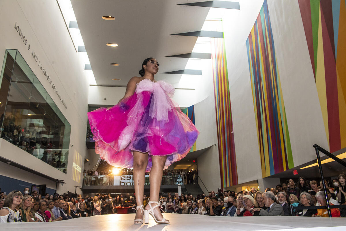A fashion model wearing a pink and purple dress with white sandals walking down a fashion runway surrounded by people at the vmfa