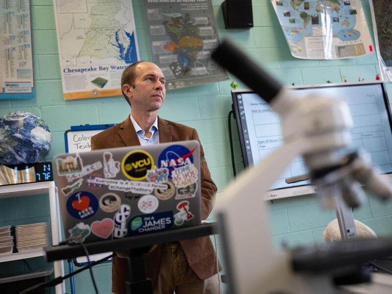 A photo of a man standing at the front of a classroom. He has a laptop in front of him covered in stickers. Behind him, the wall is covered in posters. There is a microscope in the foreground of the photo. 