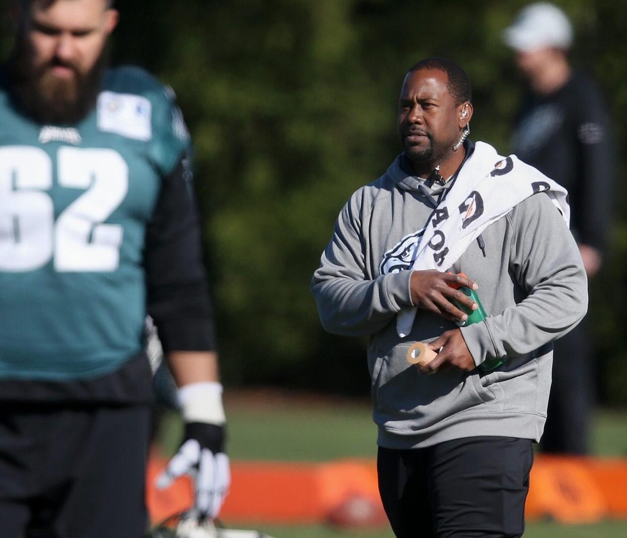Two men standing on a field. The man on the left is wearing a football uniform and the man on the right is wearing a gray hoodie with a towel over his shoulder. 