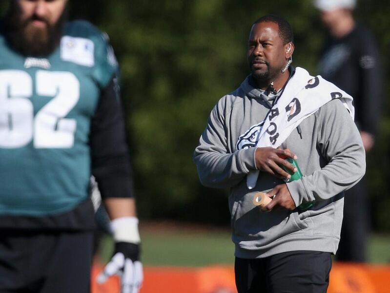 Two men standing on a field. The man on the left is wearing a football uniform and the man on the right is wearing a gray hoodie with a towel over his shoulder. 