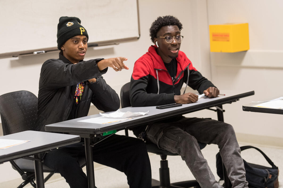 Two men sitting at a table, the man on the left is pointing at something in front of him 