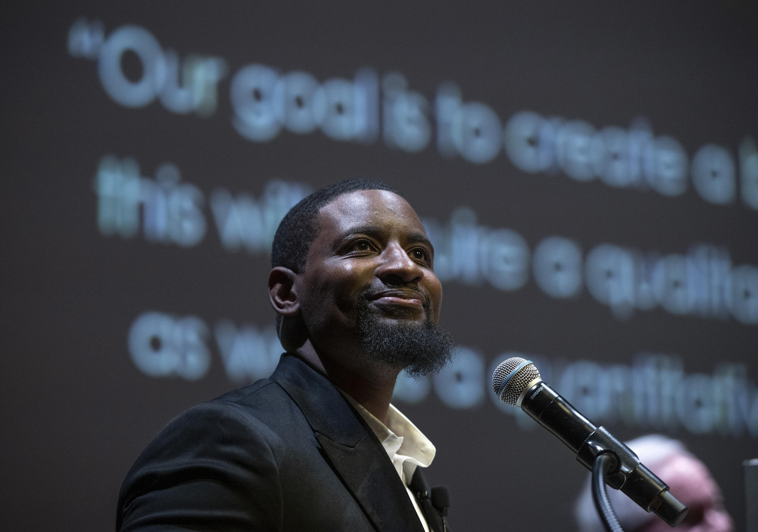 Man smiles behind a microphone and looks into an audience.