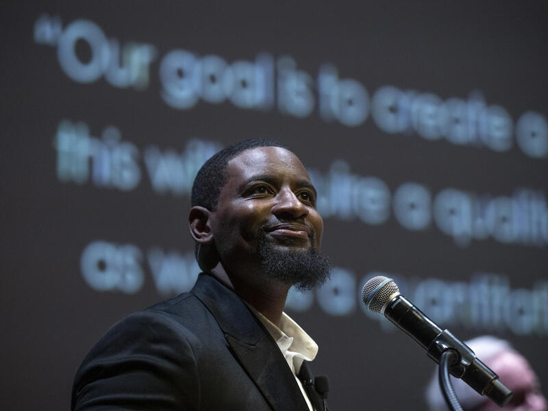 Man smiles behind a microphone and looks into an audience.