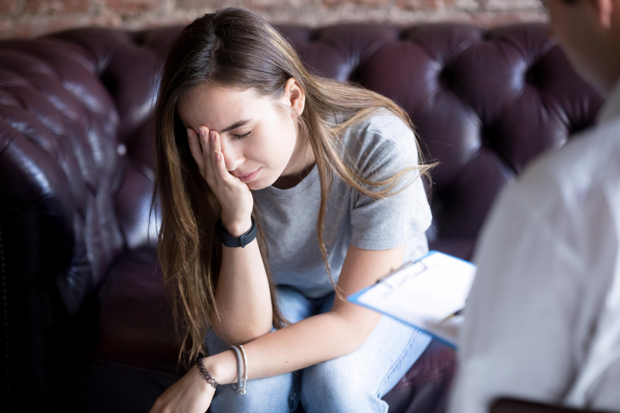 Young female appears stressed, holding her head in her hand. Someone in the foreground with a notebook appears to be communicating with her.