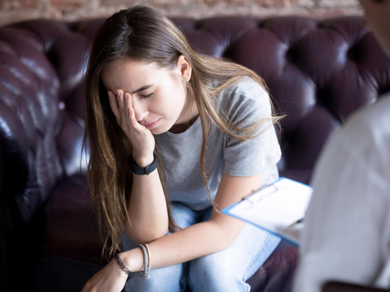 Young female appears stressed, holding her head in her hand. Someone in the foreground with a notebook appears to be communicating with her.