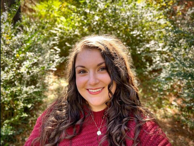 A photo of a woman standing in front of bushes. 