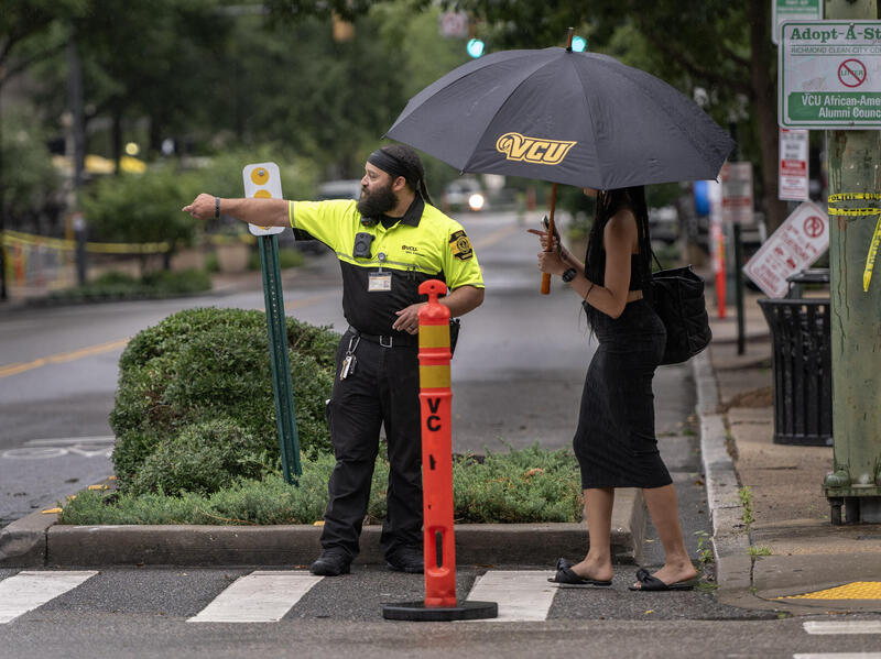 A photo of a safety officer directing a woman with an umbrella across a cross walk