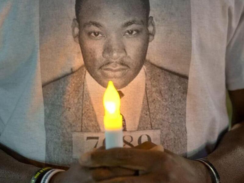 A T-shirt with Martin Luther King Jr.'s face on it. In front of the shirt are two hands holding a fake light up candle. 