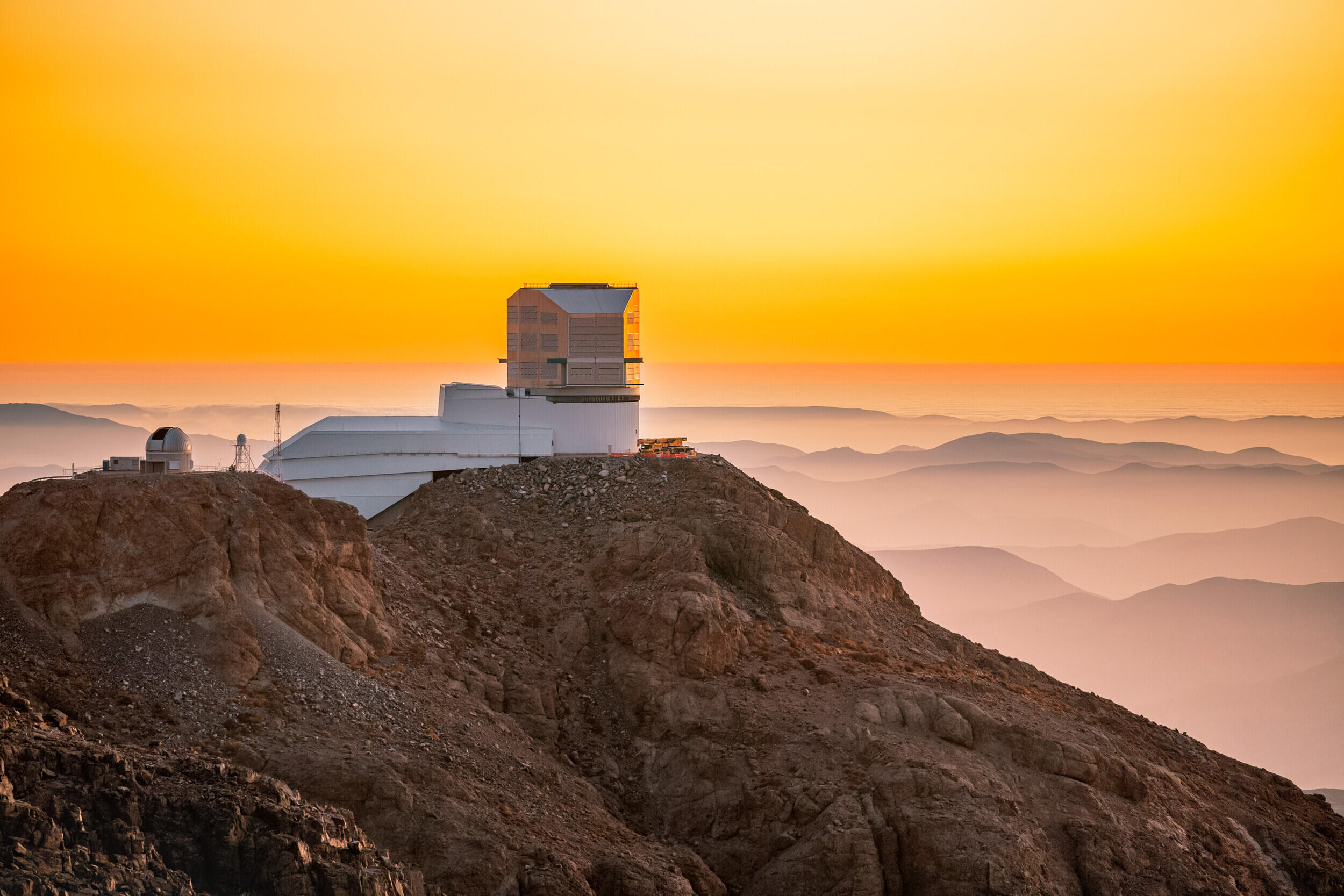 A photo of an observatory that is on a rocky mountain top. A mountain range can be seen in the background and the sky is orange because the sun is rising. 