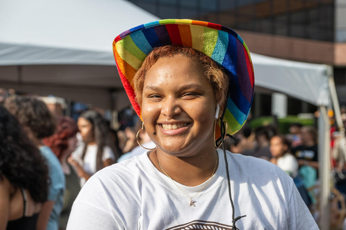 A photo of a person from the shoulders up wearing a white t-shirt and a rainbow cowboy hat. 