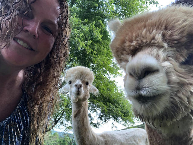 A smiling woman and two alpacas look at the camera.