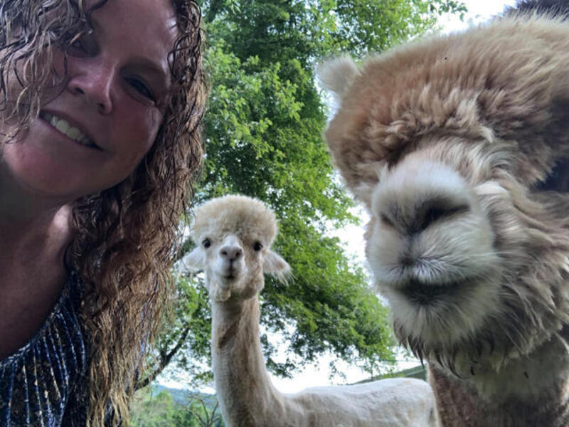 A smiling woman and two alpacas look at the camera.