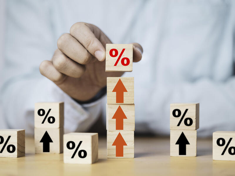 A photo of a chest and arm with the hand putting a wooden block with a percentage symbol on it on top of three other blocks with arrows pointed upwards on them. Around the table are other wooden blocks marked with upward arrows and percentage symbols. 