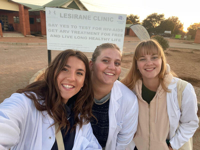 A photo of three women from the chest up smiling. They are standing in front of a sign that says \"LESIRANE CLINIC\" \"SAY YES TO TEST FOR HIV AIDS GET ARV TREATMENT FOR FREE AND LIVE LONG HEALTHY LIFE.\"