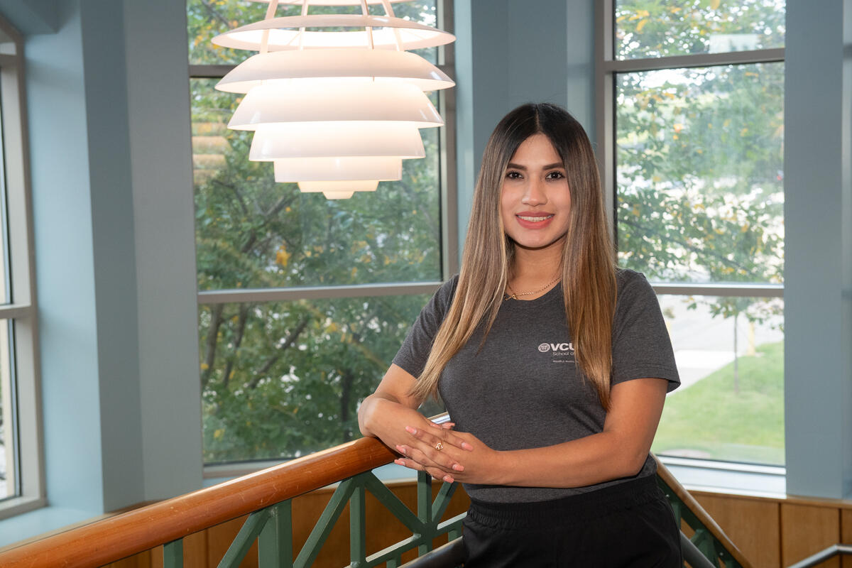 A photo of a woman from the waist up. She is leaning on the railing of the top of a staircase. 