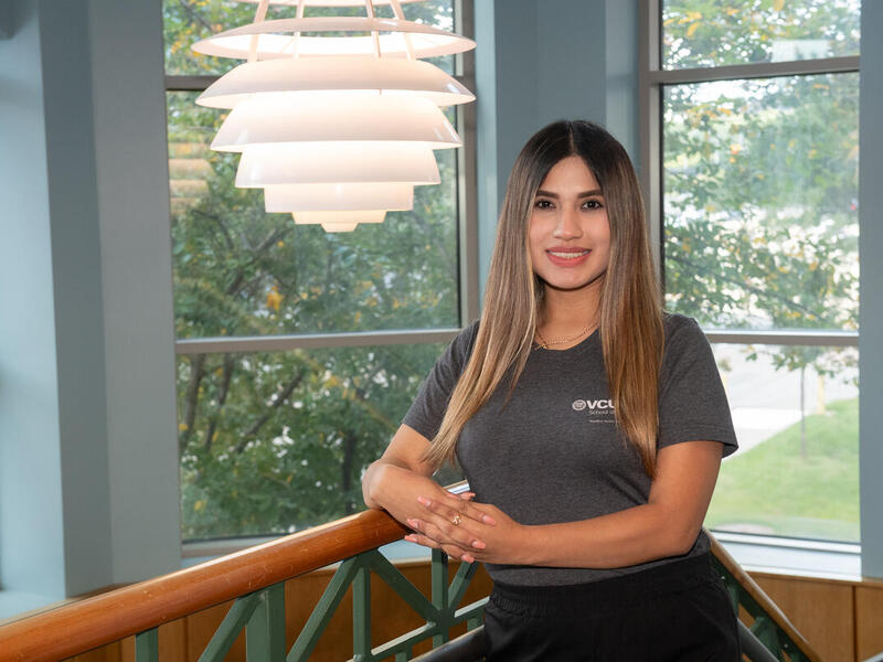 A photo of a woman from the waist up. She is leaning on the railing of the top of a staircase. 
