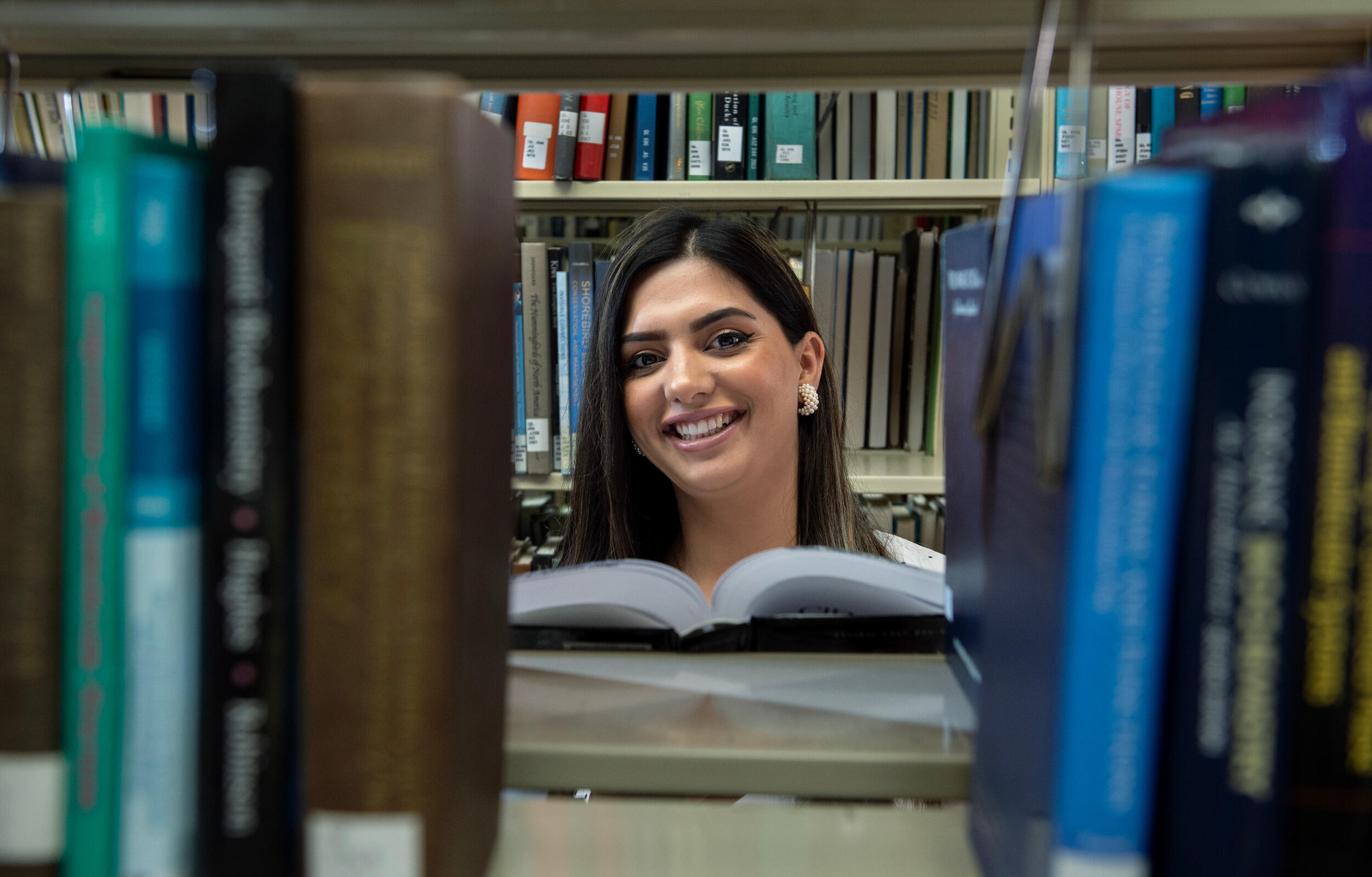 A smiling woman is seen through an opening in a shelf of books.