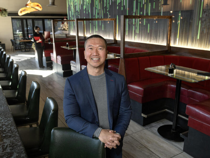 A photo of a man standing in a restaurant. He is leaning on one of the chairs along a bar. To his right are for red restaurant booths.