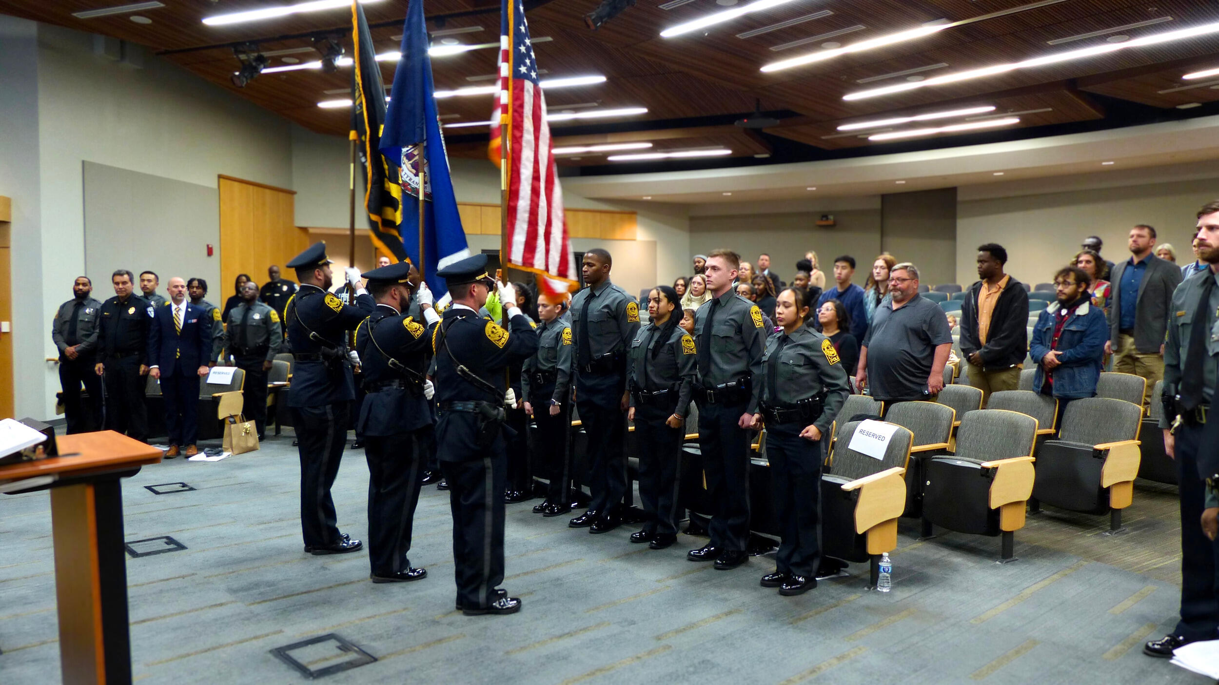 A photo of a room full of people standing. In the front are three people holding flags. 