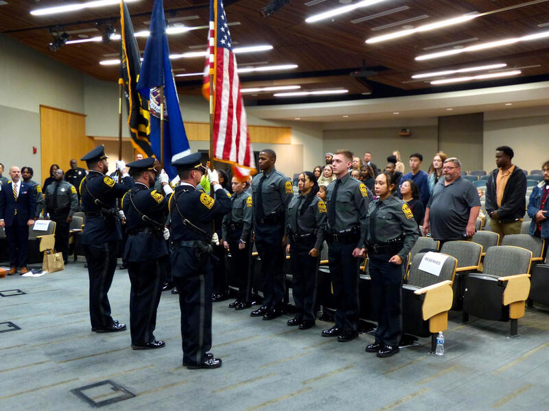 A photo of a room full of people standing. In the front are three people holding flags. 