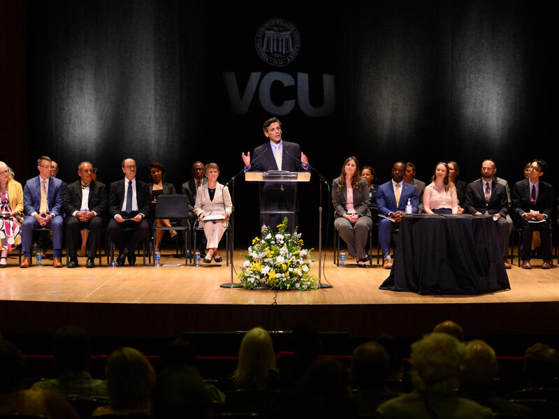 A man stands at a lectern on a stage with a group of people seated on chairs behind him.