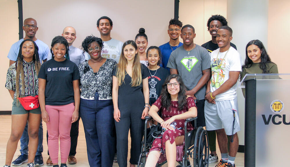 A group of people, must standing and one using a wheelchair, stand by a lectern reading V C U.