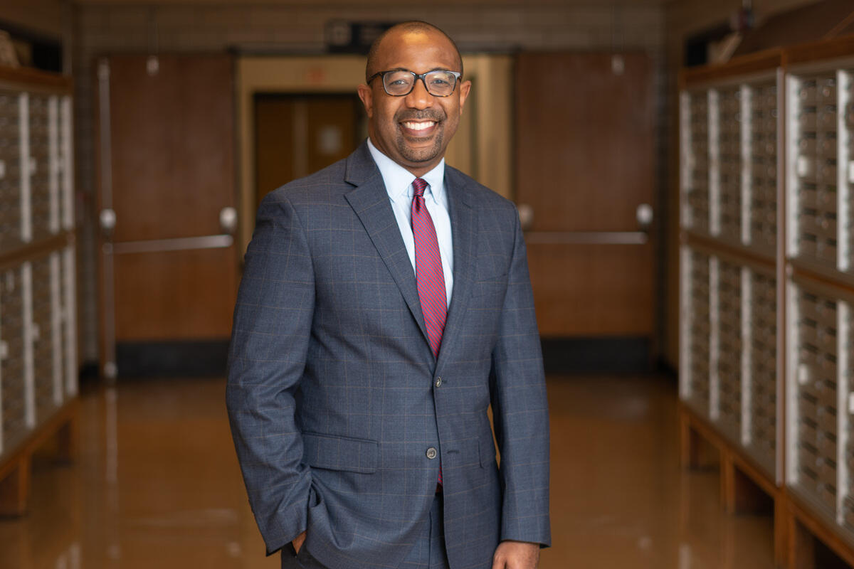 A smiling and standing in a hallway. He's wearing a gray suit, white button up shirt, a maroon tie, and glasses. 