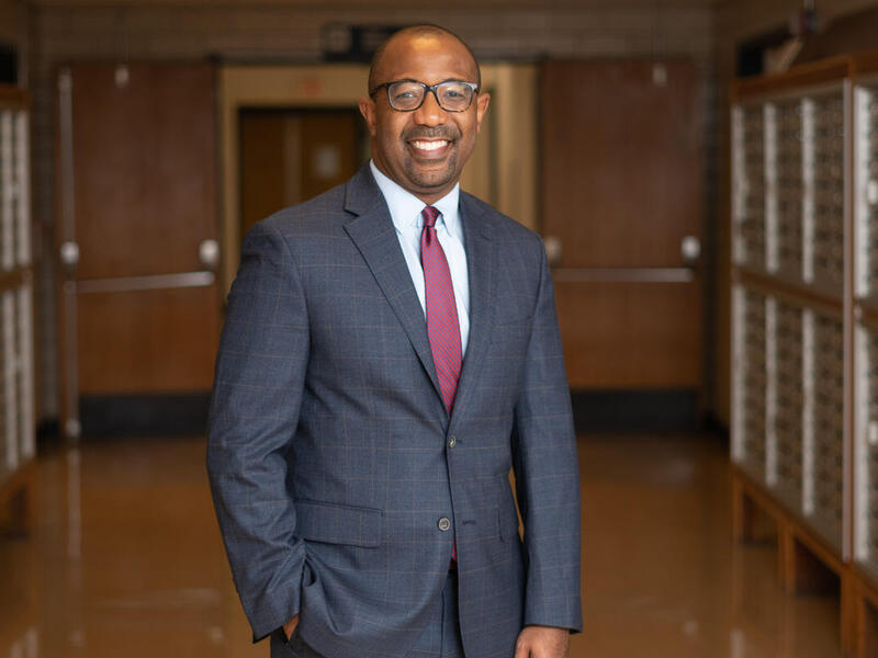 A smiling and standing in a hallway. He's wearing a gray suit, white button up shirt, a maroon tie, and glasses. 