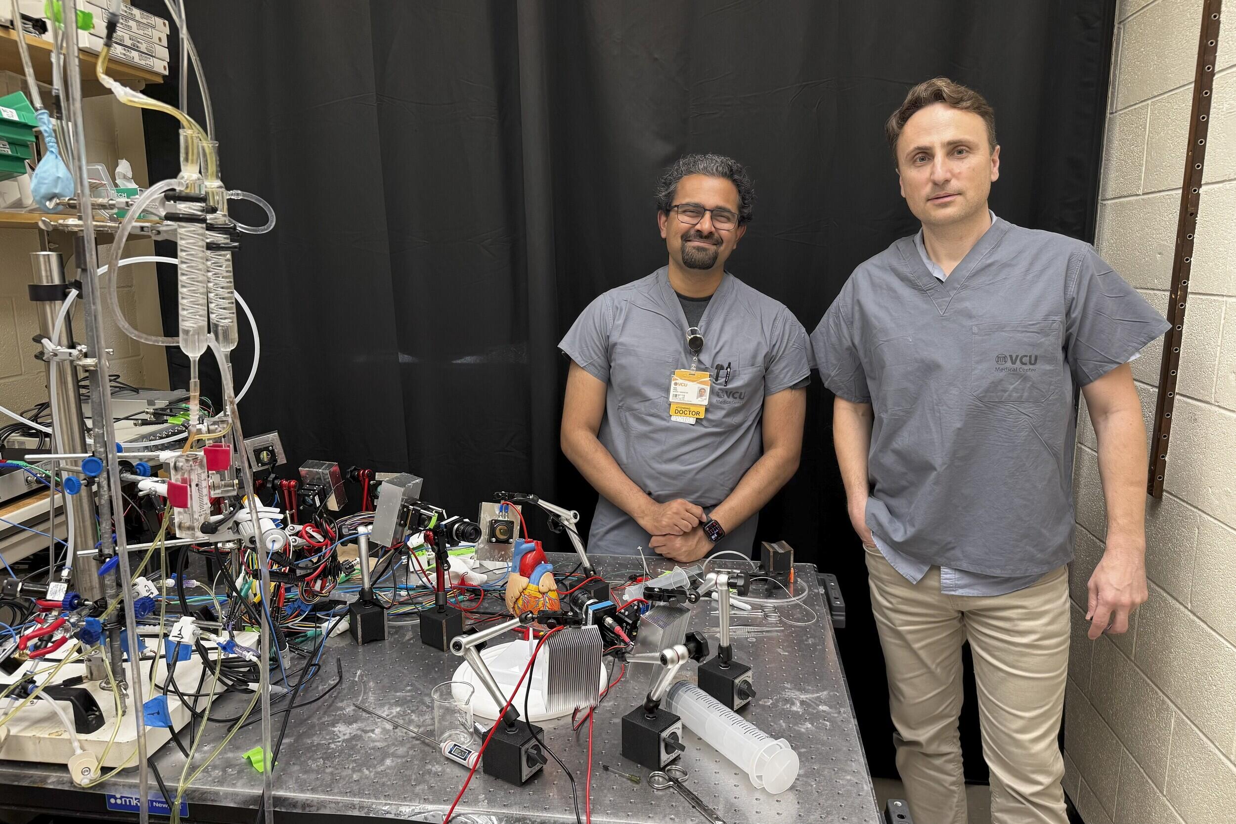 A photo of two men standing behind a table covered in wires and pieces of equipment. 