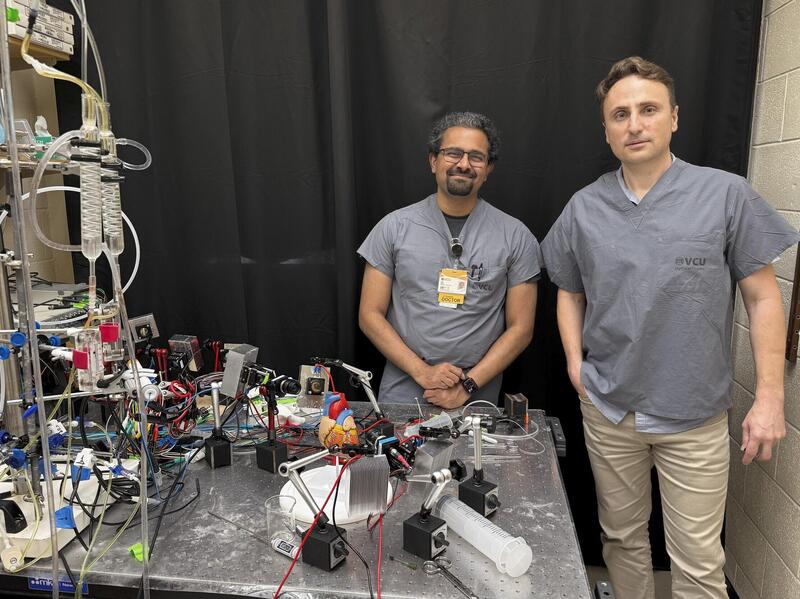 A photo of two men standing behind a table covered in wires and pieces of equipment. 