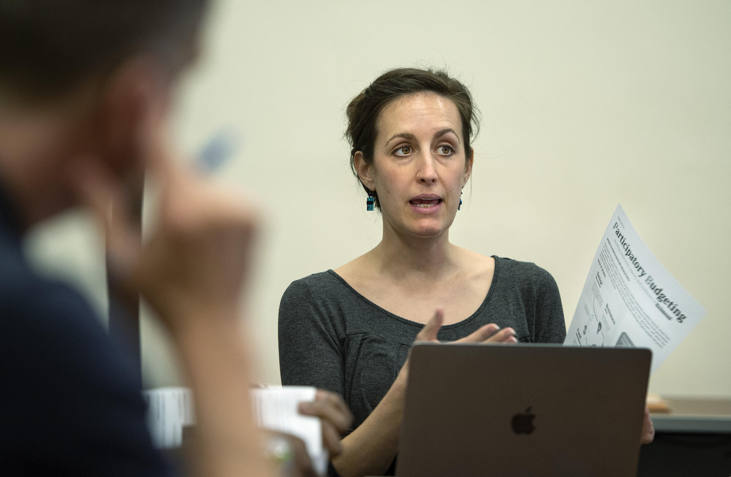 A woman speakingshile sitting at a table. She is gesturing to a piece of paper in her right hand. 