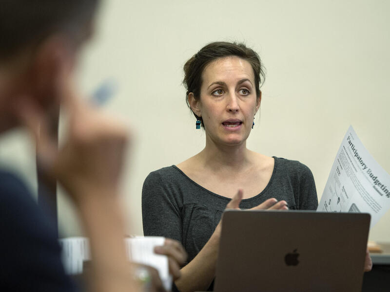 A woman speakingshile sitting at a table. She is gesturing to a piece of paper in her right hand. 