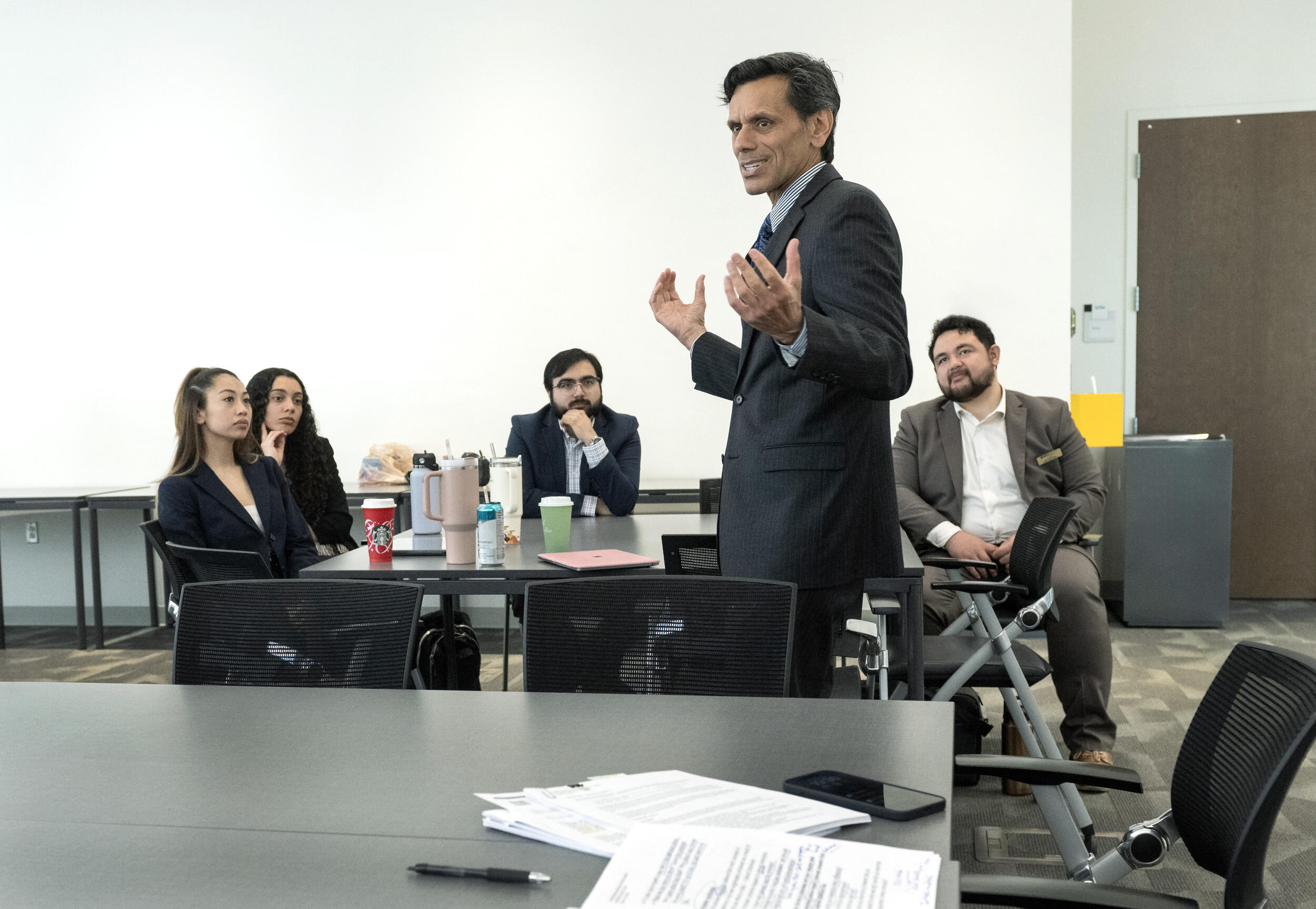 A photo of a man standing in between two tables and talking. The table behind the man has four people sitting at it while watching him speak. 