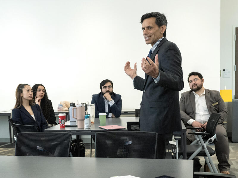 A photo of a man standing in between two tables and talking. The table behind the man has four people sitting at it while watching him speak. 
