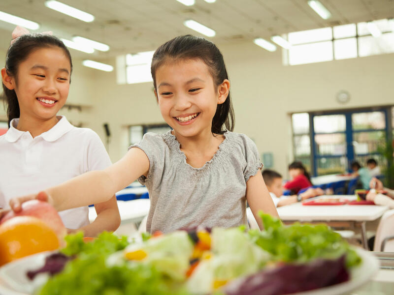 Two young girls smile and select food in a school cafeteria-type environment.