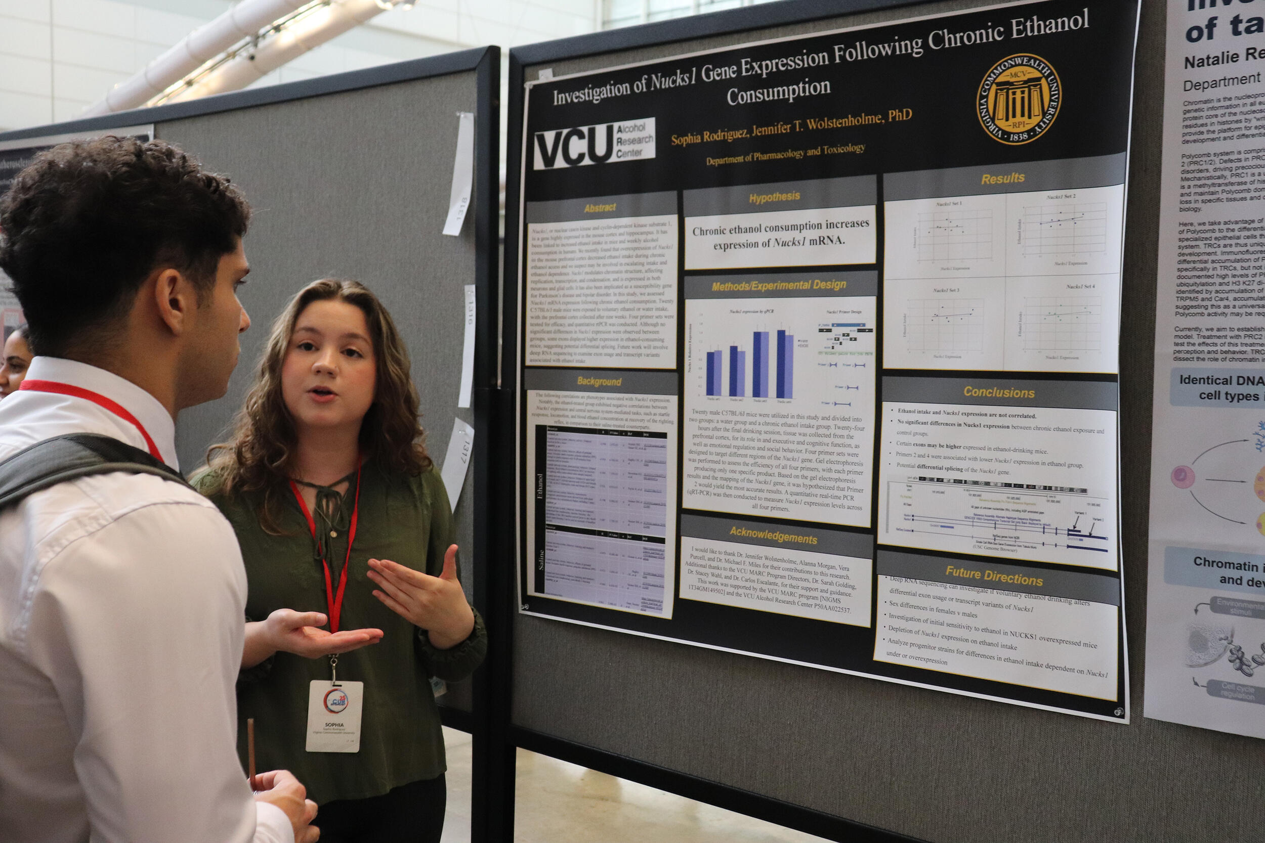 A photo of a woman and man standing to the right of a research poster. The woman is speaking and the man is listening to her. 